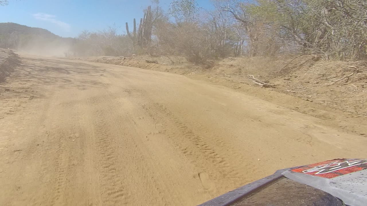 Sandy road curving through scrub with dust ahead and vehicle hood at the bottom edge.
