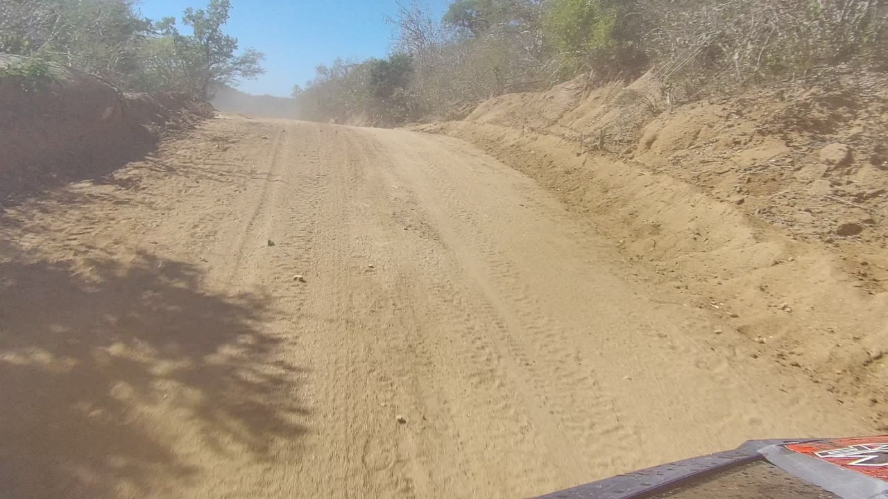 Straight sandy road with scrub banks and a vehicle edge in the foreground.