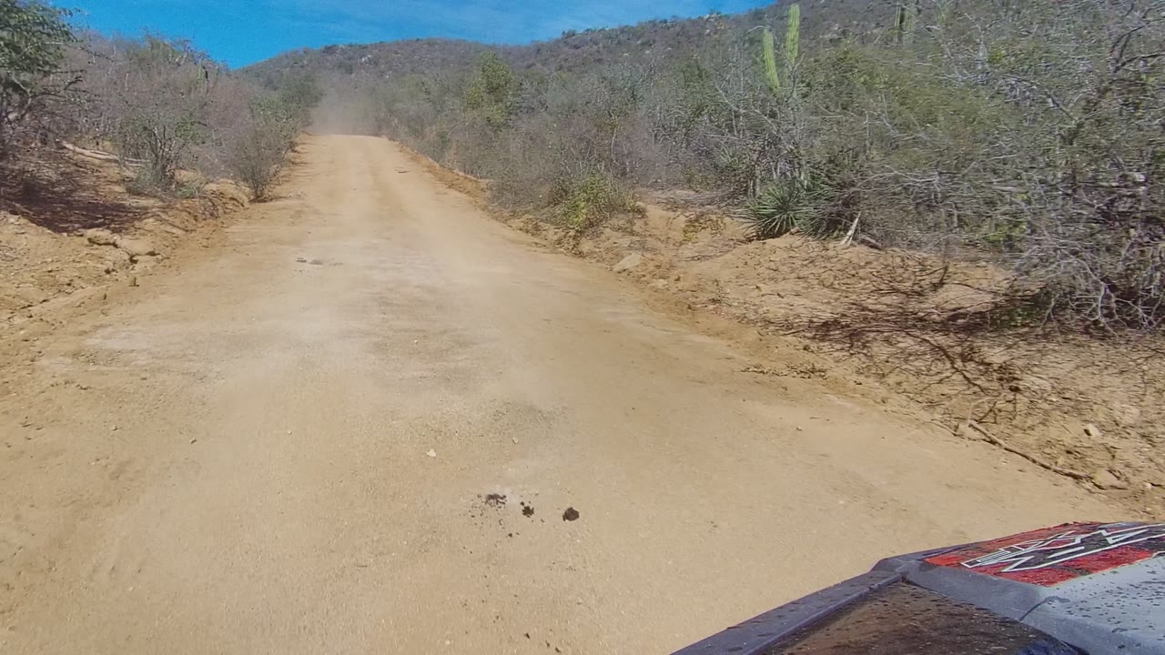 Forward view of a sandy dirt road in Los Cabos, Baja California Sur, with dry scrub on both sides, low hills ahead, a vehicle hood edge at bottom right, and no visible people.