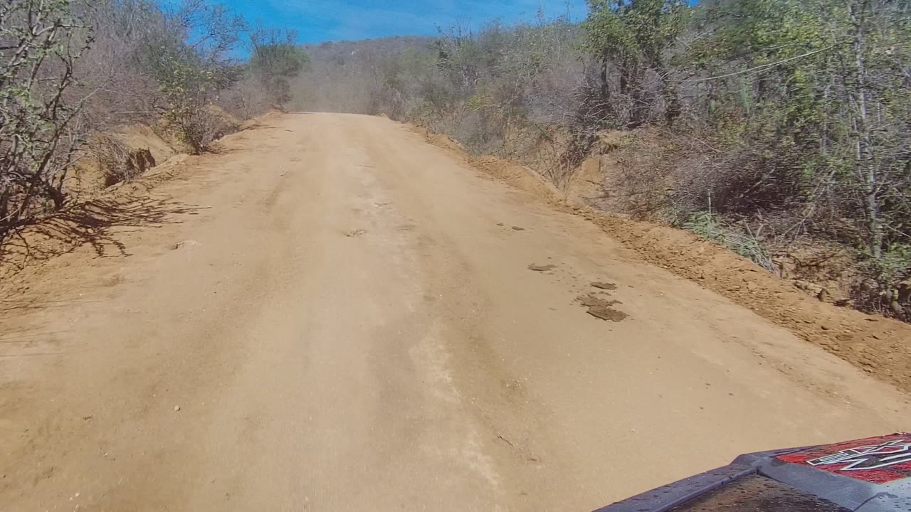 The Los Cabos dirt road appears wider in frame, with uneven sandy surface, brushy raised shoulders, distant low hills, clear blue sky, and no visible people.