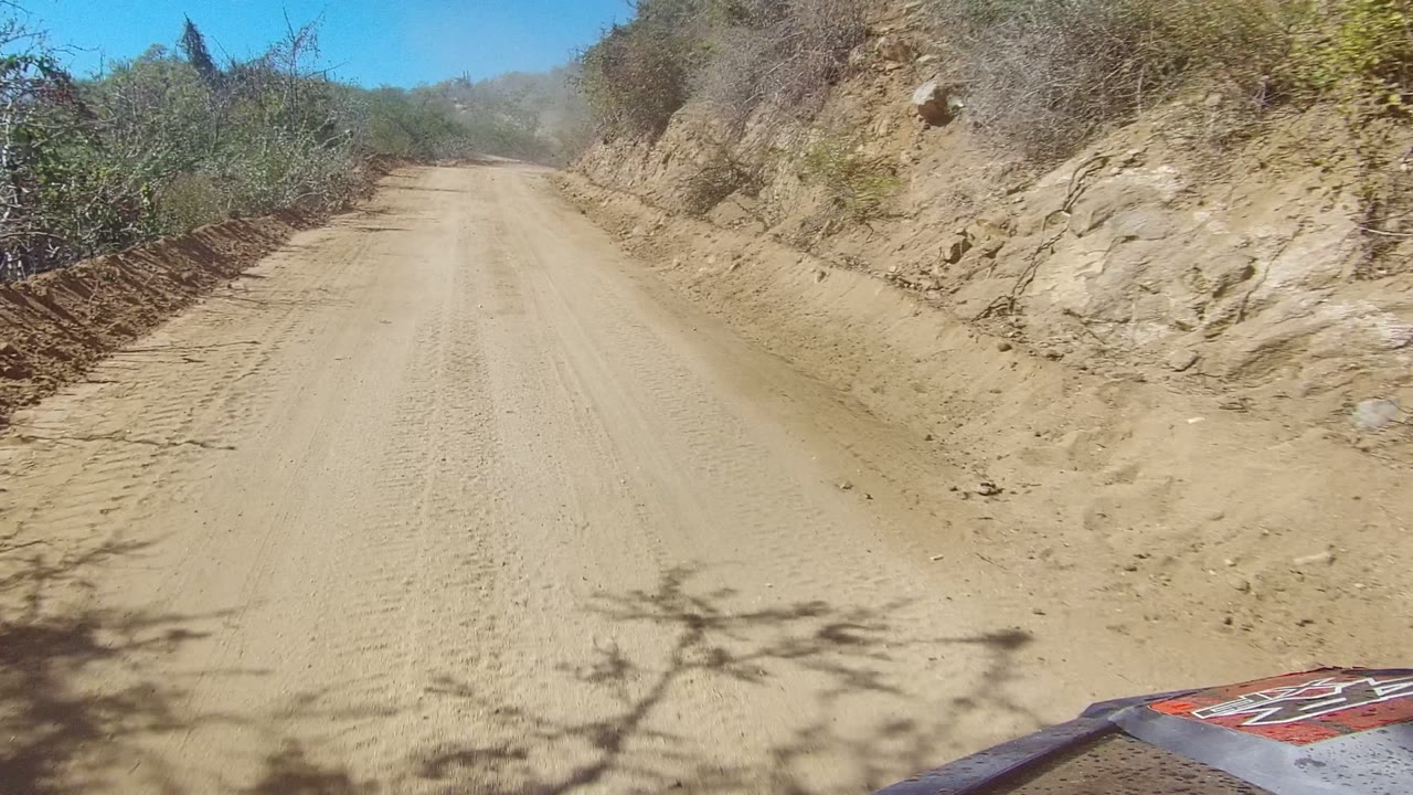 Low front-vehicle view of a dusty dirt road in Los Cabos, Baja California Sur, with dry brush on both sides, rocky tan banks, blue sky, and no visible people.