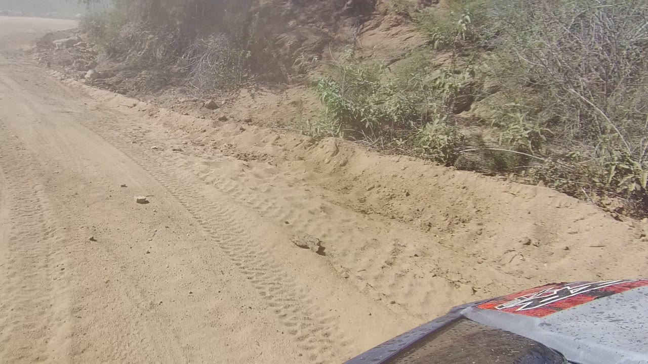 Dust hangs over the sandy road ahead in Los Cabos, Baja California Sur, with rocky roadside edges, sparse shrubs, and no visible people.