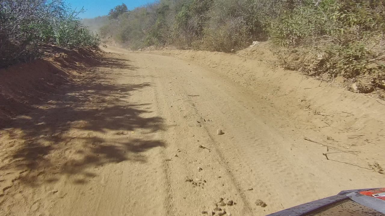 A sandy Los Cabos dirt track continues through dry roadside vegetation, with stronger shadow patches and a partial vehicle edge visible at lower right, no visible people.