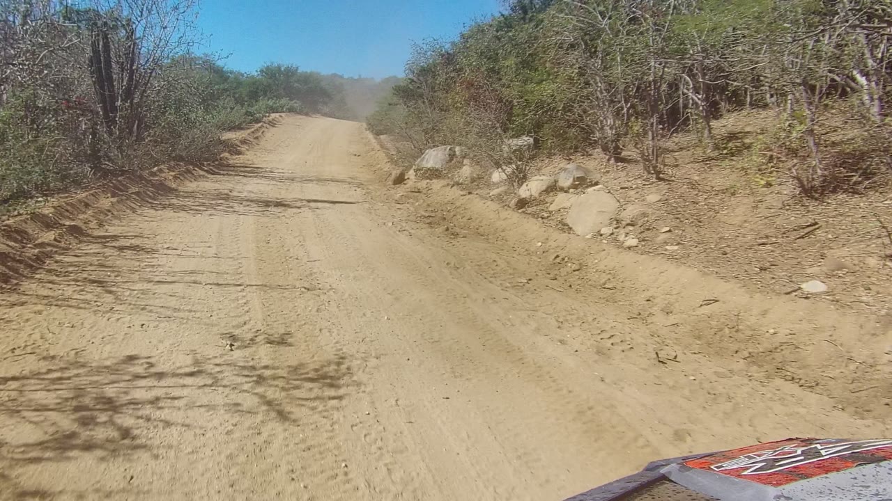 Forward vehicle view on a Los Cabos sandy road, with scrubby brush, rocks on the right shoulder, light dust ahead, and no visible people.