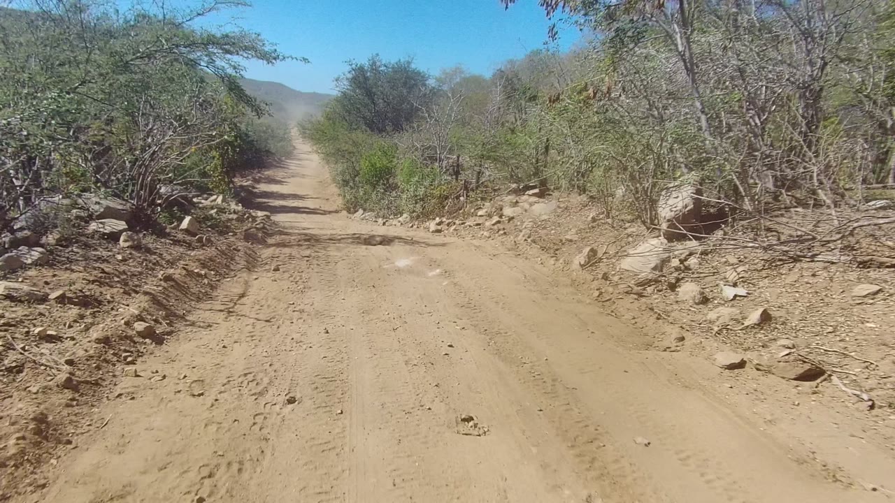 Opening view of a sandy dirt road in Los Cabos, Baja California Sur, with dry shrubs, roadside rocks, distant hills, and no visible people.