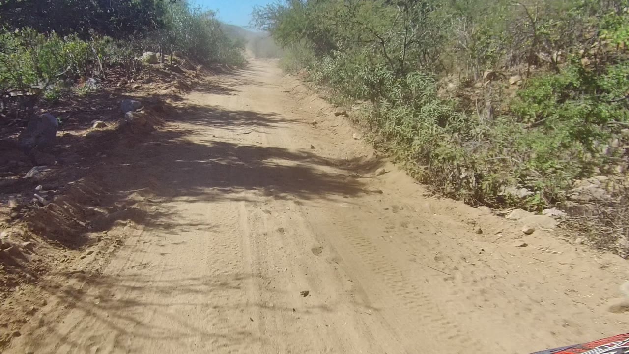 Mid-clip view of the Los Cabos dirt track with stronger shadow patterns on the sand, dry roadside vegetation, and no visible people.