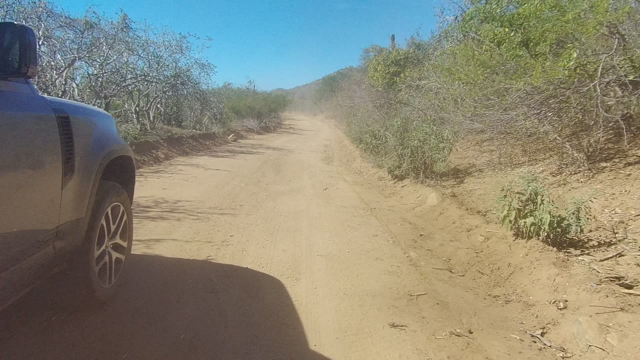 Early frame of a sandy dirt road in Los Cabos, Baja California Sur, with dry roadside brush, a partial vehicle edge at left, dust ahead, and no visible people.