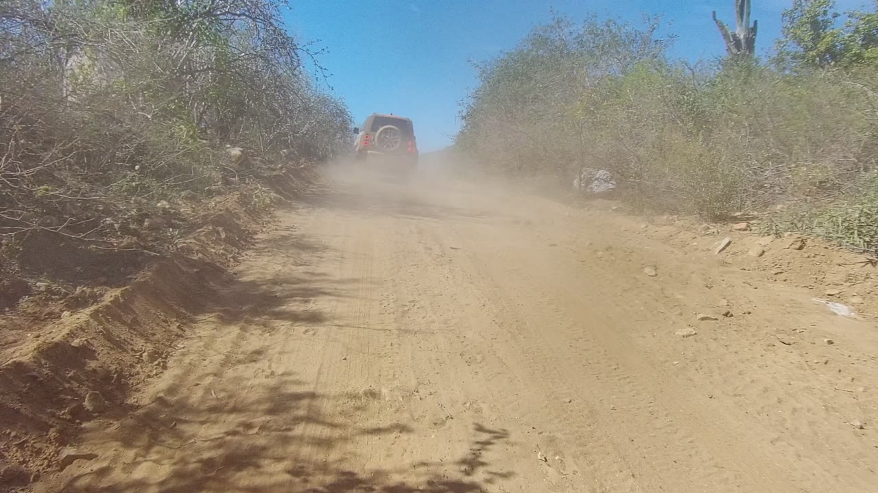 A dusty sandy road in Los Cabos, Mexico, with an SUV near the upper left moving away between dry roadside brush and no visible people.