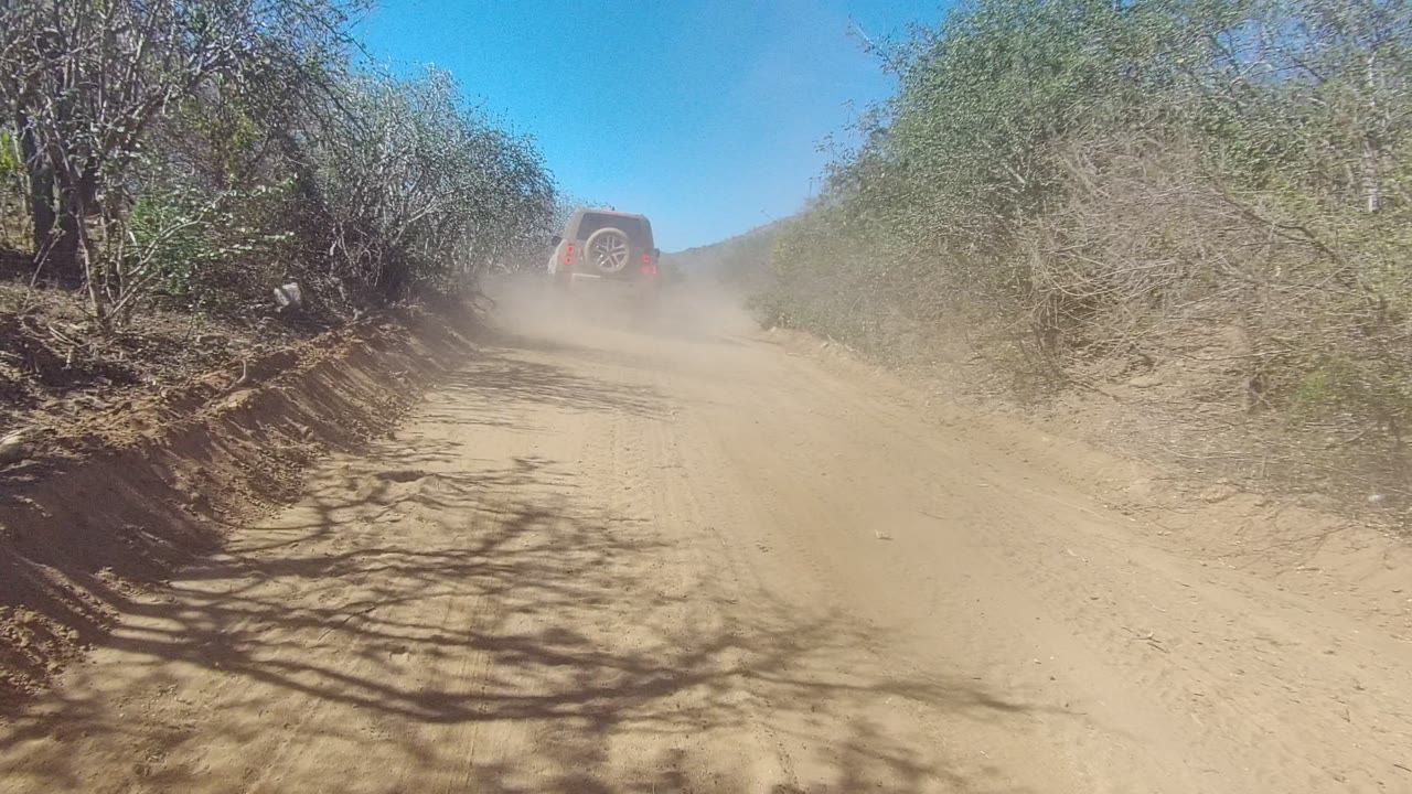 The SUV remains distant through dust on a sandy Los Cabos road, with tree shadows crossing the track, dry brush on both sides, and no visible people.