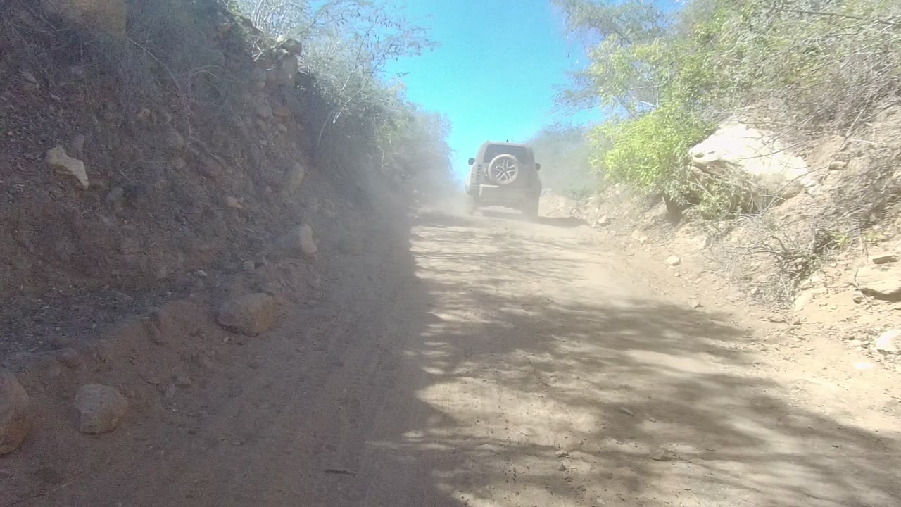 A dark SUV drives away on a dusty dirt road in Los Cabos, Baja California Sur, with rocky banks, scrub vegetation, blue sky, and no visible people.
