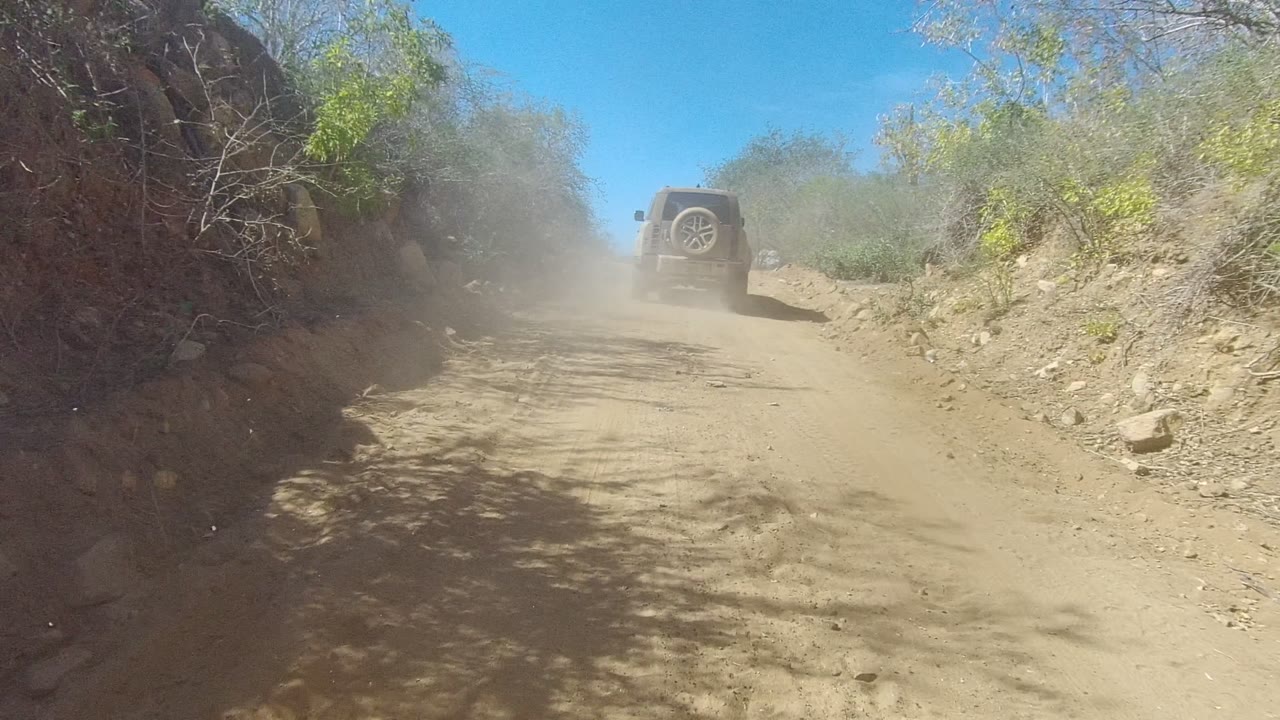 A dusty Los Cabos off-road track curves slightly upward toward a dark SUV, with dry vegetation, rocky edges, tree shadows across the road, and no visible people.