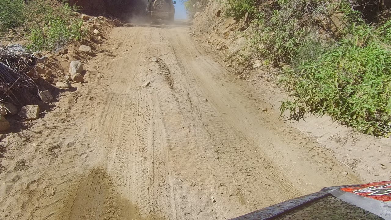 A dusty narrow dirt road in Los Cabos, Baja California Sur climbs between rocky slopes and sparse brush, with a vehicle ahead near the top and no visible people.