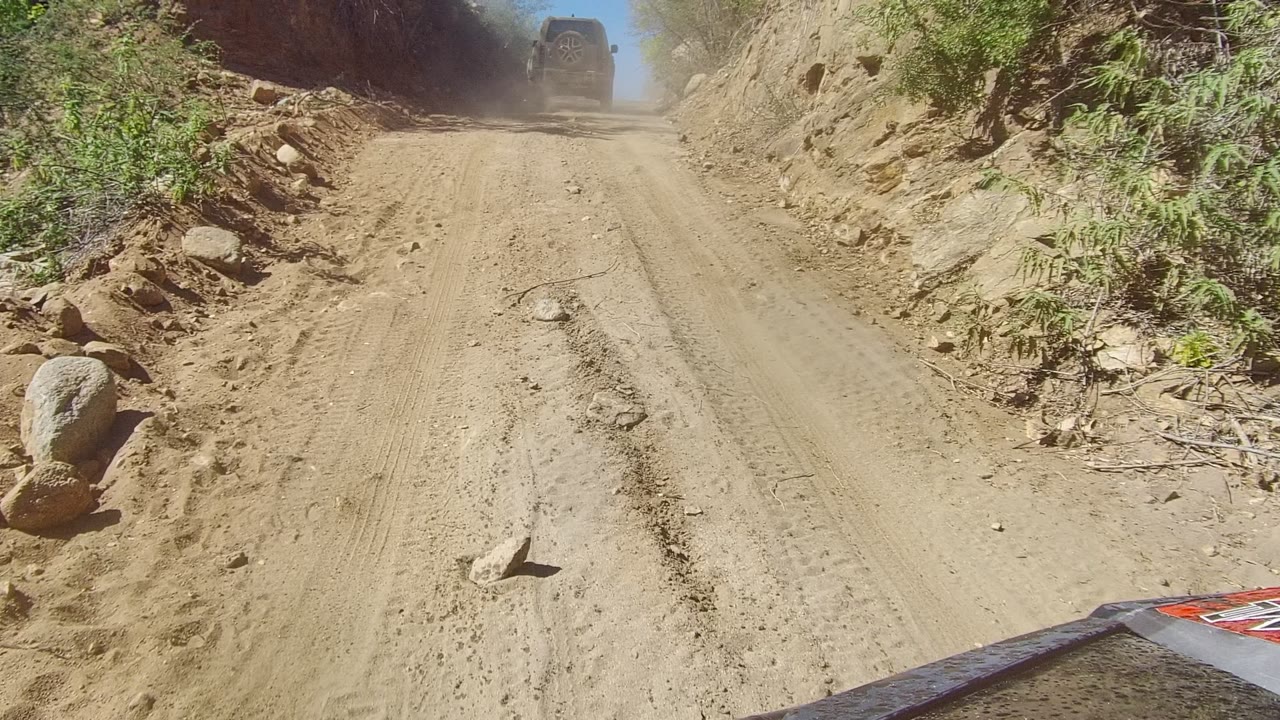 The road remains centered as a lead vehicle moves farther through a dusty cut in the rocky Los Cabos terrain, with dry plants on the left and no visible people.