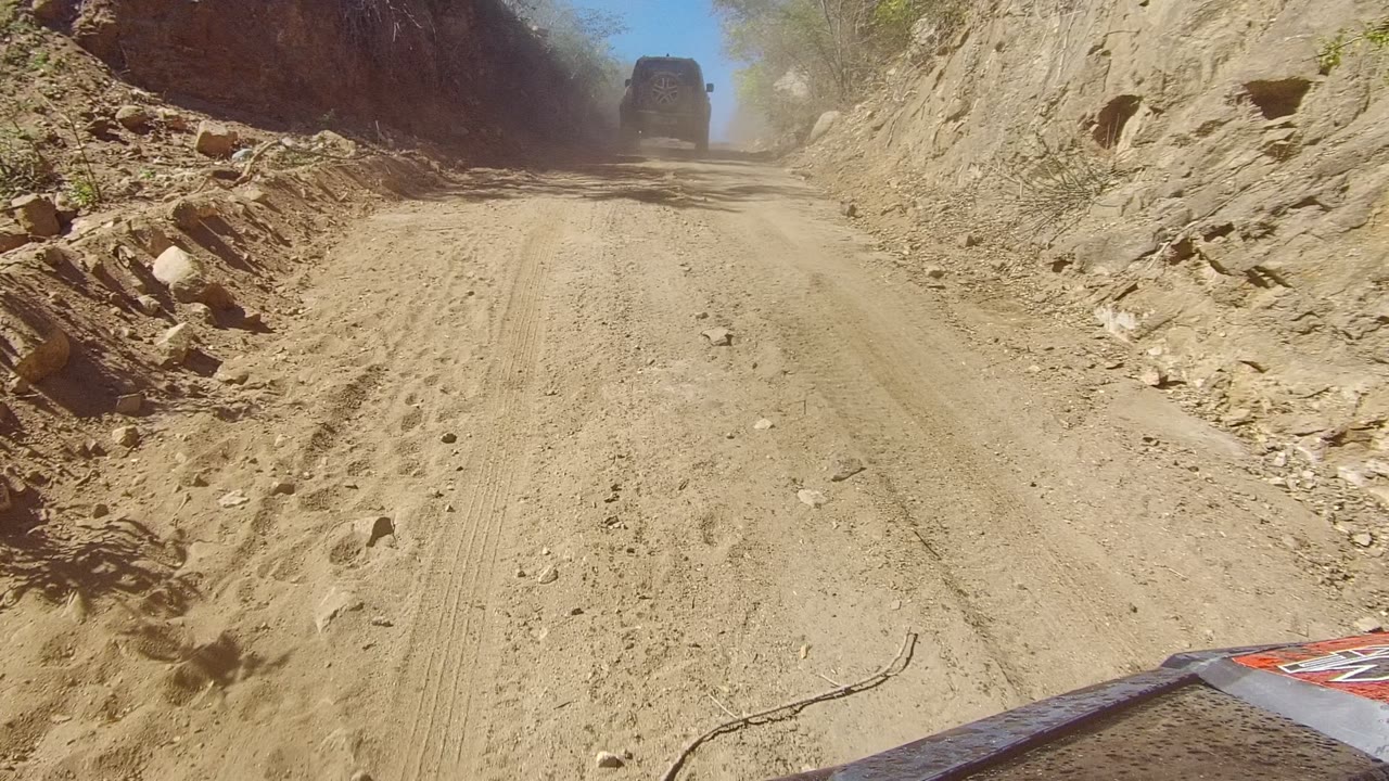 A lead vehicle is partly obscured by dust near the upper right of a narrow tan road bordered by eroded rock walls and scattered brush in Los Cabos, with no visible people.