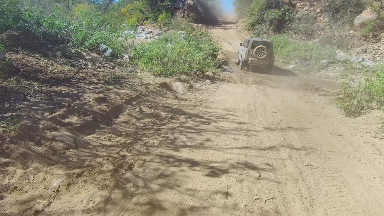 A light-colored 4x4 drives away on a muddy trail in Los Cabos, Baja California Sur, with green brush, rocks, and no visible people.