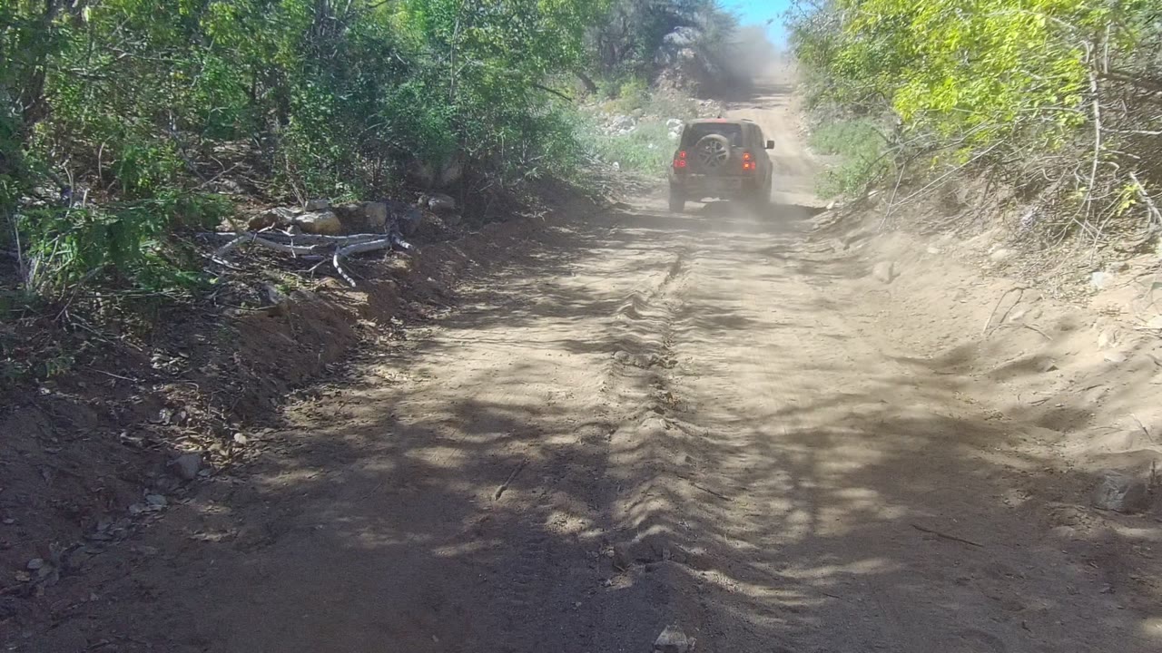 A distant SUV climbs the uneven dirt road in Los Cabos, with darker shade across the foreground, bright dust ahead, roadside rocks, and no visible people.