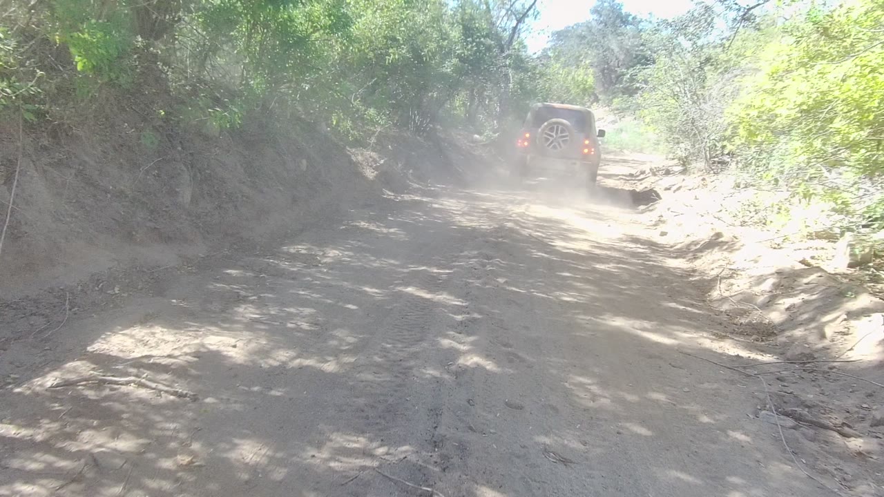 The SUV continues away along the uneven Los Cabos dirt road, with dust softening the background and dense green brush tight along both sides, no visible people.
