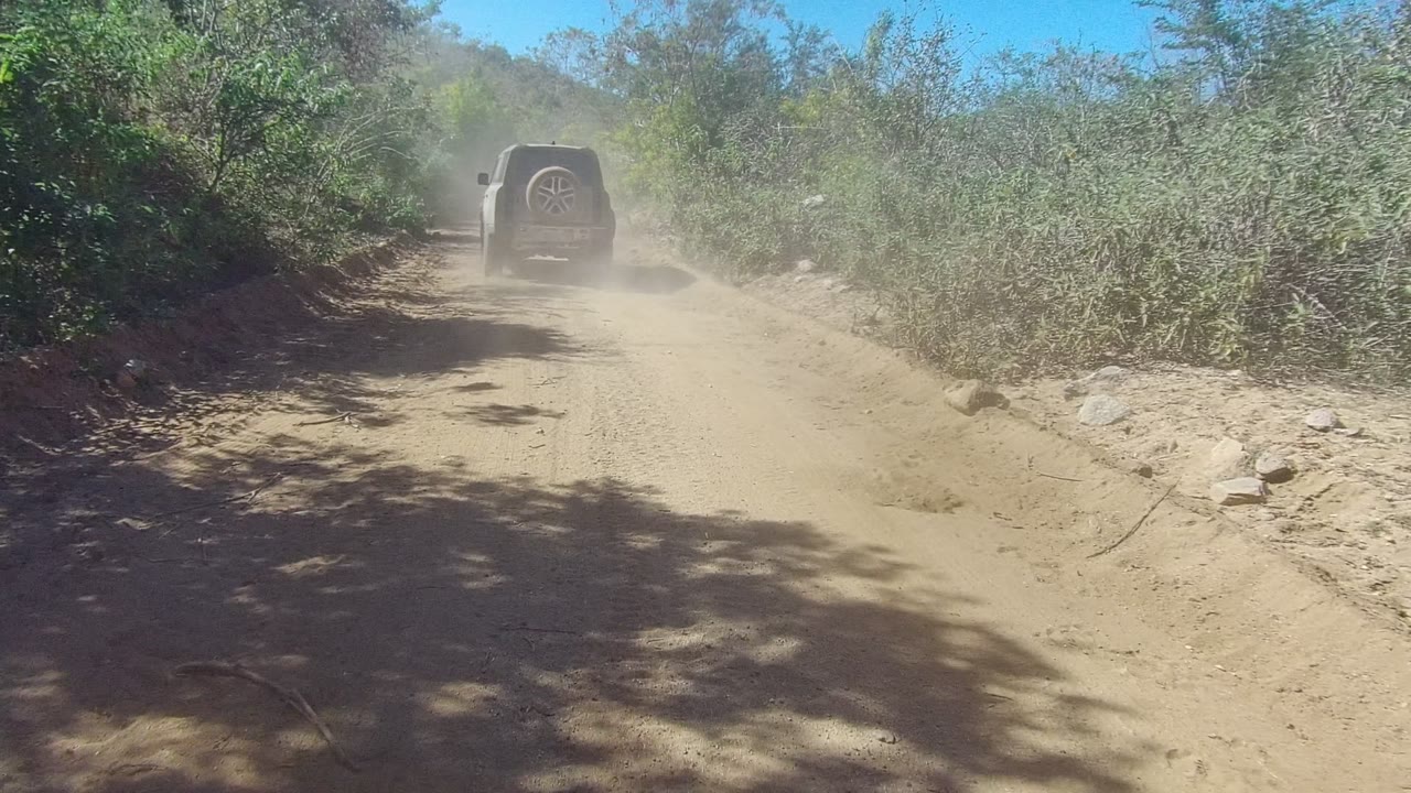 At 0:00 in Los Cabos, a dark SUV drives away on a narrow tan dirt road with green bushes on both sides, dust rising behind it, and no visible people.