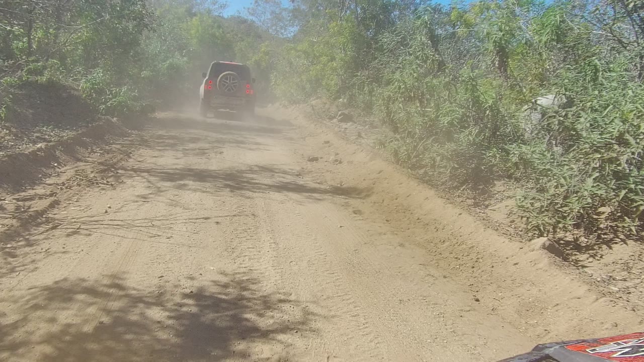 At 0:02 in Los Cabos, the SUV moves toward the upper right of the road as dust spreads across the trail, with brush banks, tree shadows, and no visible people.