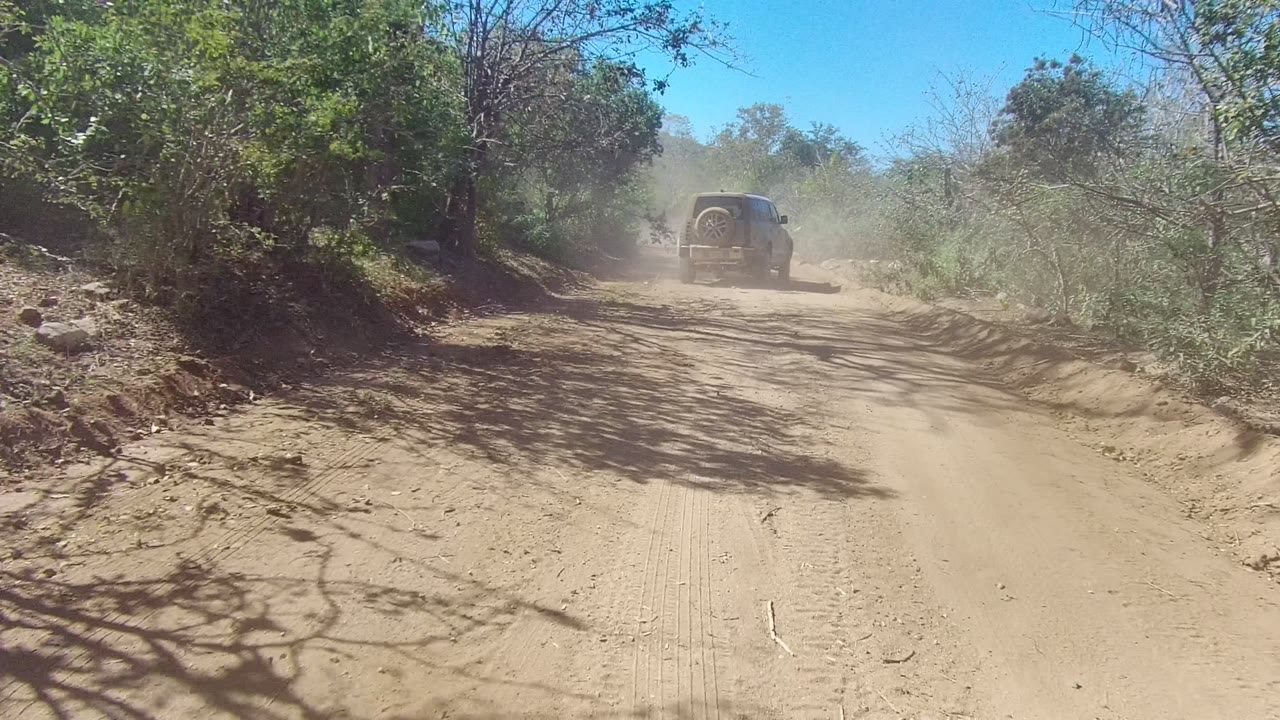 The SUV continues away near the right side of the Los Cabos dirt road, bordered by scrubby green vegetation and dry branches, no visible people.