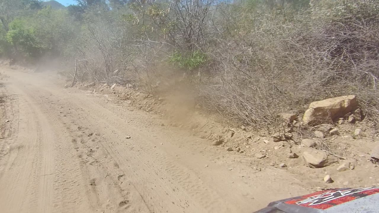 A dusty dirt road in Los Cabos, Baja California Sur curves past dry brush and scattered rocks, with a vehicle hood edge at the bottom and no visible people.
