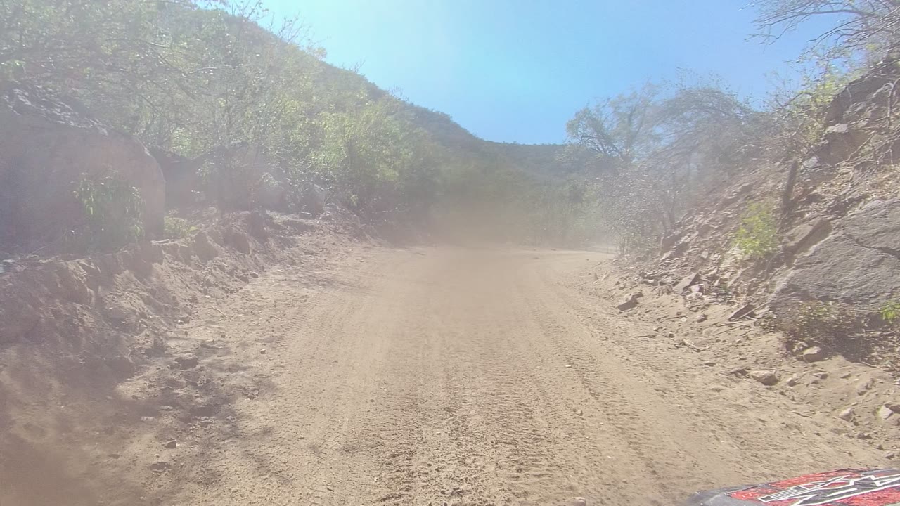 A dusty dirt road in Los Cabos, Baja California Sur curves past rocky roadside slopes and dry scrub under a clear blue sky, with no visible people.