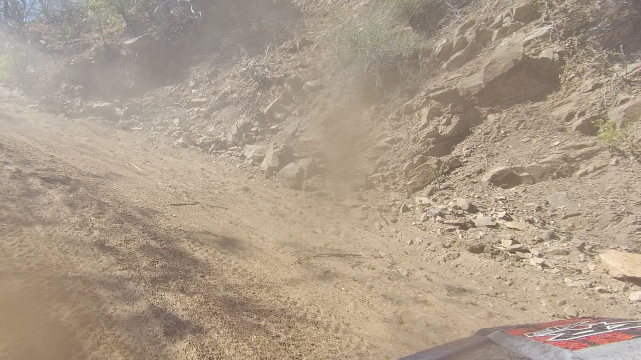 Forward view on a dusty mountain dirt road in Los Cabos, Baja California Sur, with rocky slope at right, sparse brush, and no visible people.