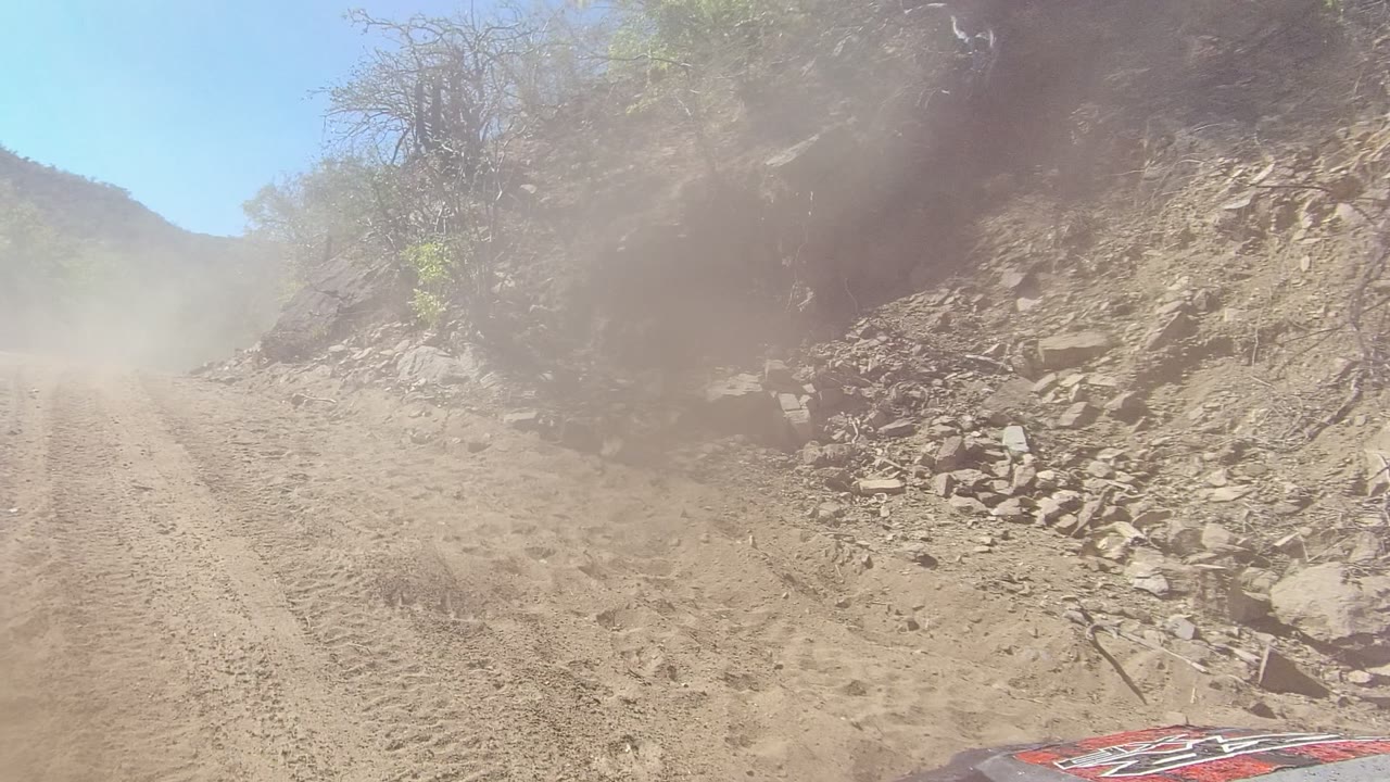 A dusty Los Cabos mountain road stretches ahead under bright sky, with a raised dust plume, rocky embankments, sparse shrubs, and no visible people.