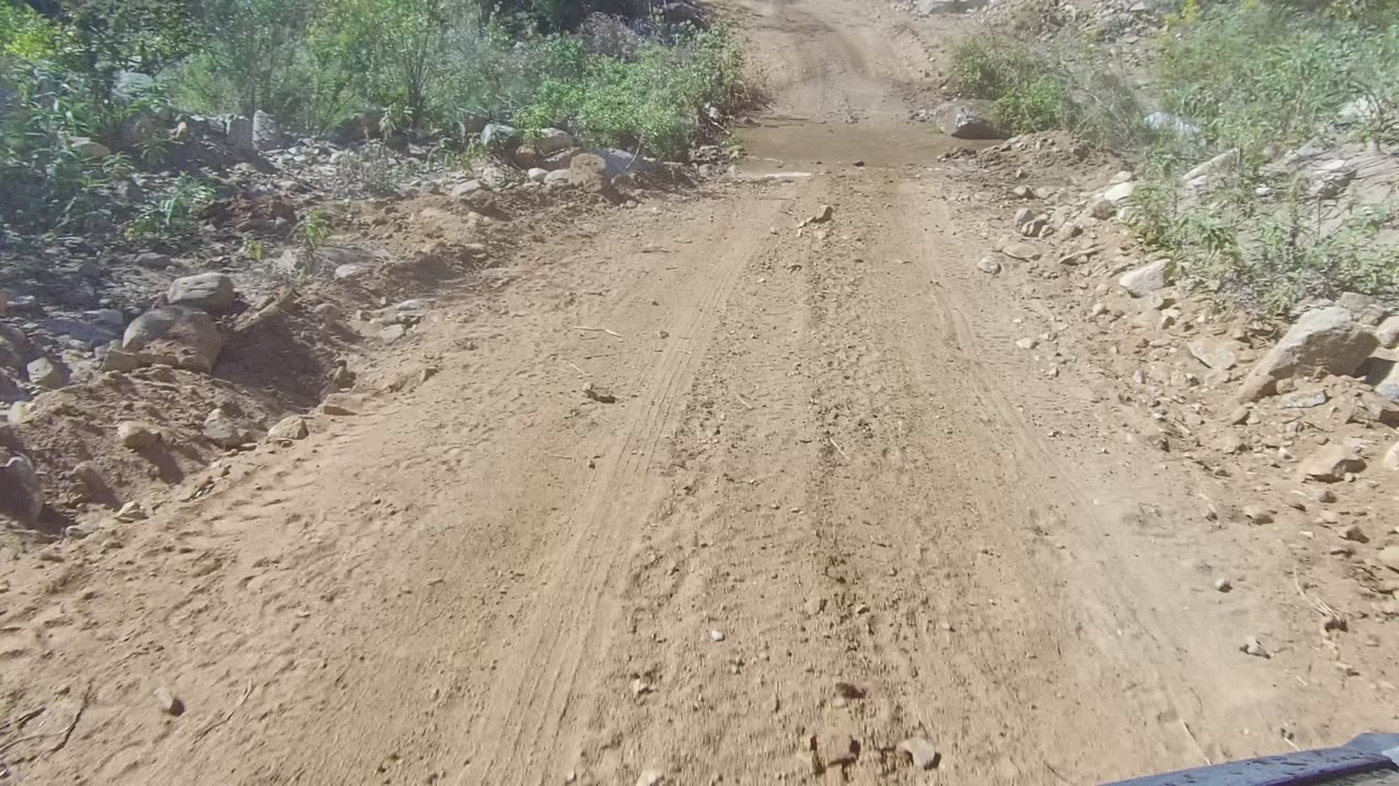A muddy unpaved road in Los Cabos, Baja California Sur stretches ahead between rocks and sparse green vegetation, with no visible people.