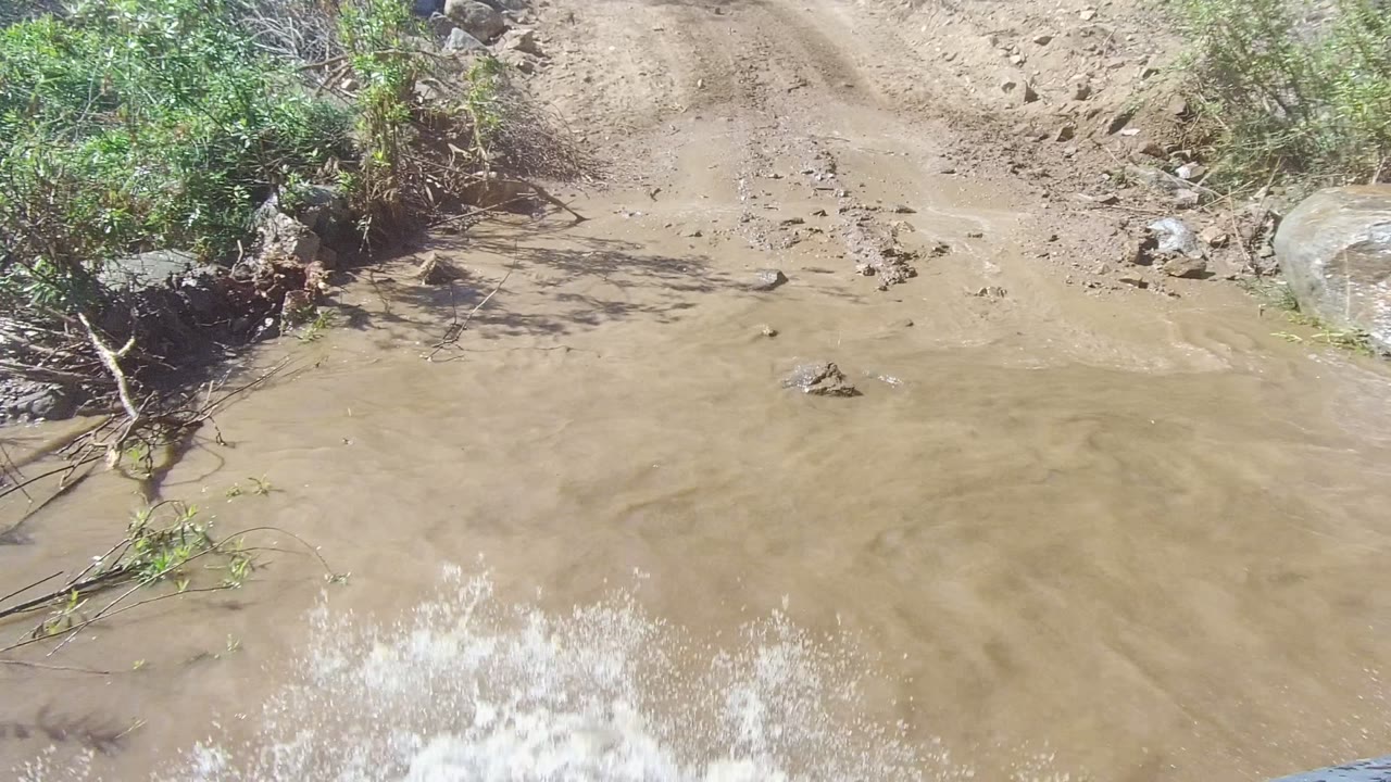 Brown water splashes across the lower right of the frame as the vehicle crosses the puddled dirt road in Los Cabos, Baja California Sur, with brush and rocks along the edge and no visible people.