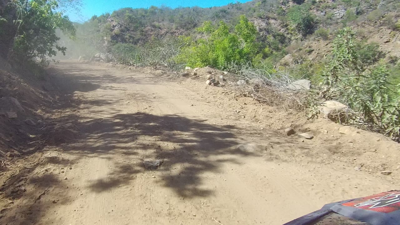 Dusty dirt road in Los Cabos, Baja California Sur, with scrub vegetation, rocky slopes, bright sun, a vehicle hood edge at lower right, and no visible people.
