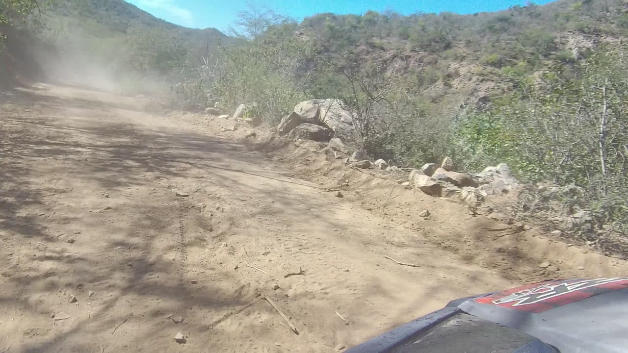 Rutted Los Cabos dirt road passing a rocky hillside, with brush on both sides, tree shadows across the track, dust ahead, and no visible people.