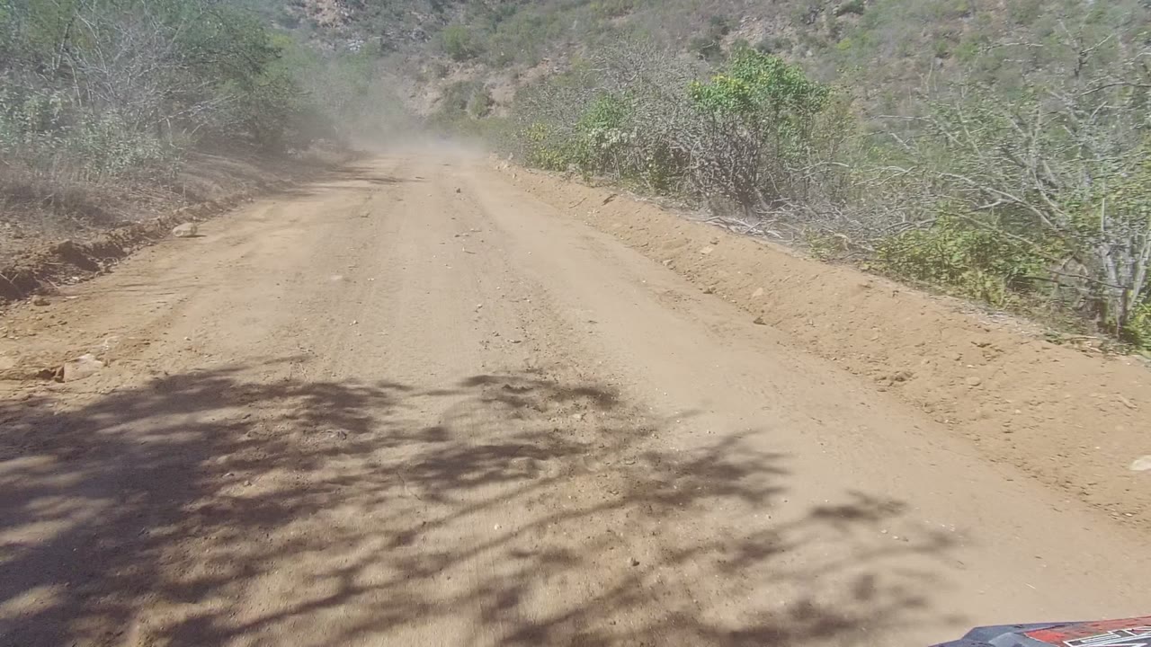 A dusty tan dirt road runs through dry brush in Los Cabos, Baja California Sur, with a dust cloud ahead and no visible people.