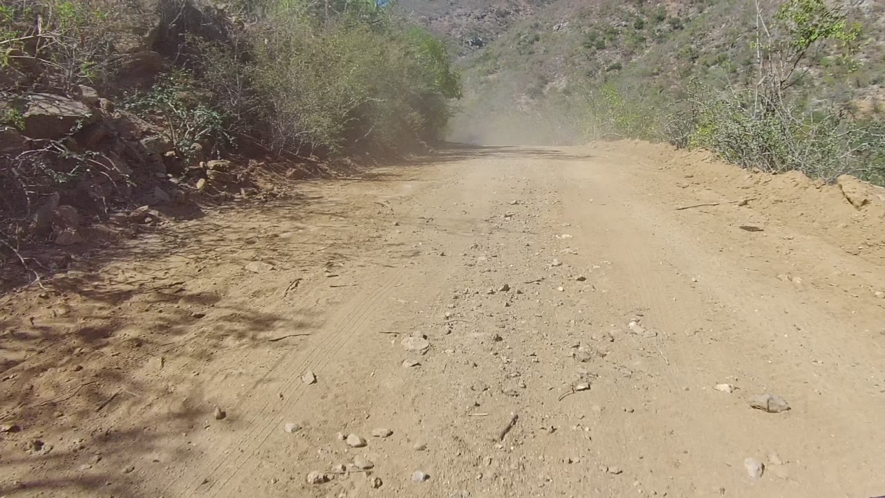 The road widens in frame as it passes a rocky edge and arid vegetation in Los Cabos, Baja California Sur, Mexico, with dust visible near the far bend and no visible people.