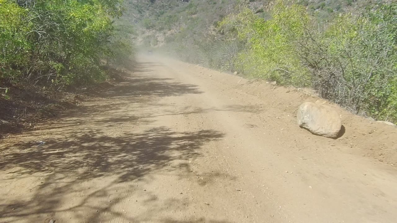 A dusty Los Cabos road passes a large rock on the right edge, with tree shadows across the tan surface and no visible people.