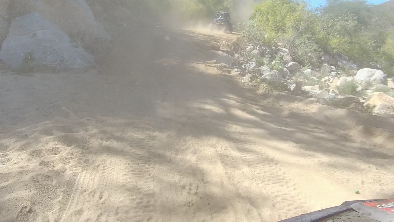 At the start of the Los Cabos clip, a dusty dirt road curves through rocky terrain with green brush on both sides and a small vehicle ahead, with no visible people.