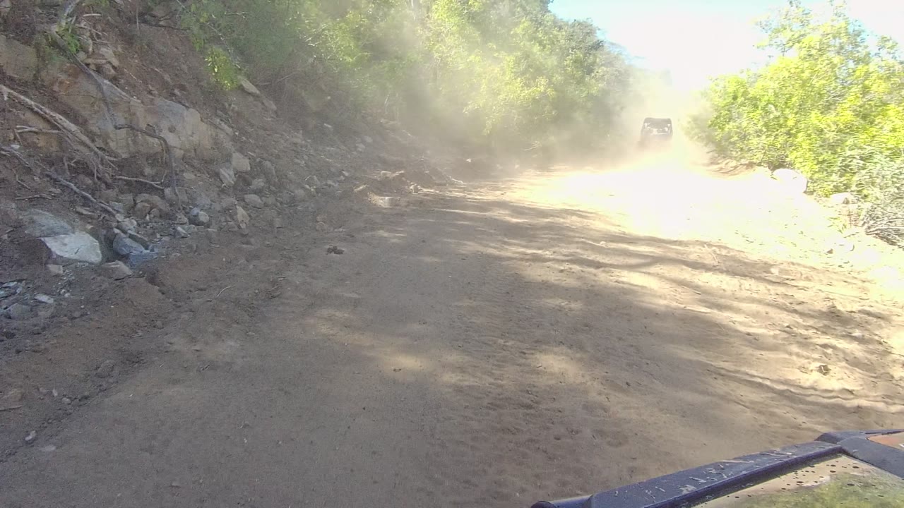 At 0:02, the road runs between dense green vegetation and a rocky bank while dust softens the background where the lead vehicle is barely visible, with no visible people.