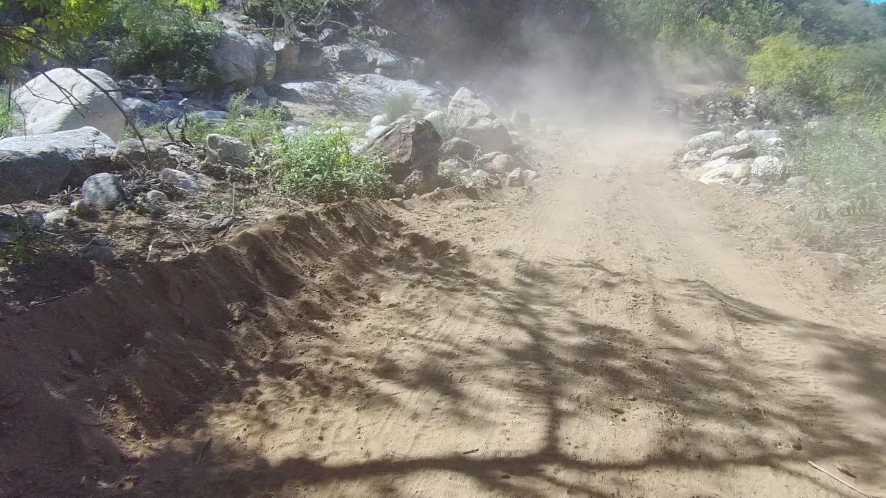 Rocky dirt trail in Los Cabos, Baja California Sur with a dust cloud hanging over the sandy road, scrub vegetation at the edges, and no visible people.