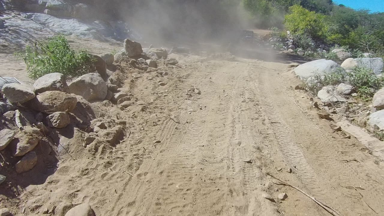 The sandy off-road track in Los Cabos, Baja California Sur passes a pile of rocks as dust spreads across the upper frame, with no visible people.