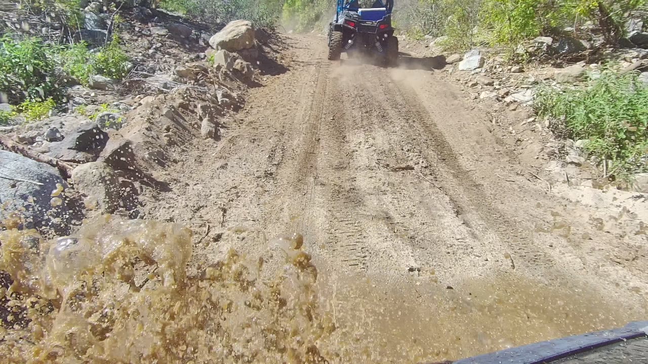 A 4x4 off-road vehicle drives away through muddy water on a rutted Los Cabos dirt road, with rocks and green vegetation at the sides and no visible people.