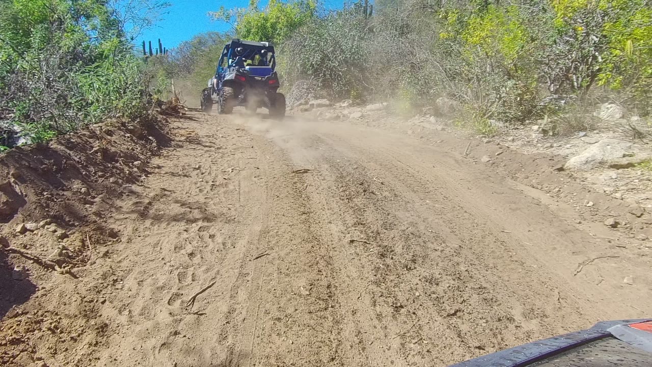 A 4x4 off-road vehicle moves farther ahead on a dry Los Cabos dirt road, raising a tan dust cloud between roadside brush and rocks, with no visible people.