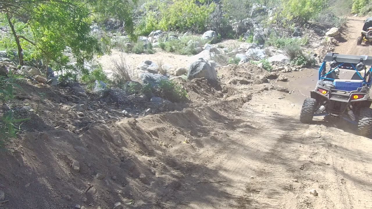 Blue ATV enters a rutted dirt trail in Los Cabos, Baja California Sur, with rocks, bushes, bright sun, and no visible people.