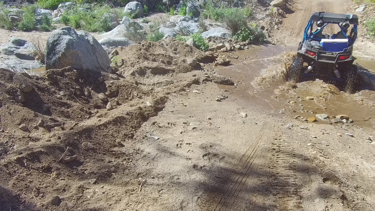 The ATV climbs along the right side of a muddy rutted trail in Los Cabos, with a large boulder, scattered rocks, and dry brush visible, and no visible people.