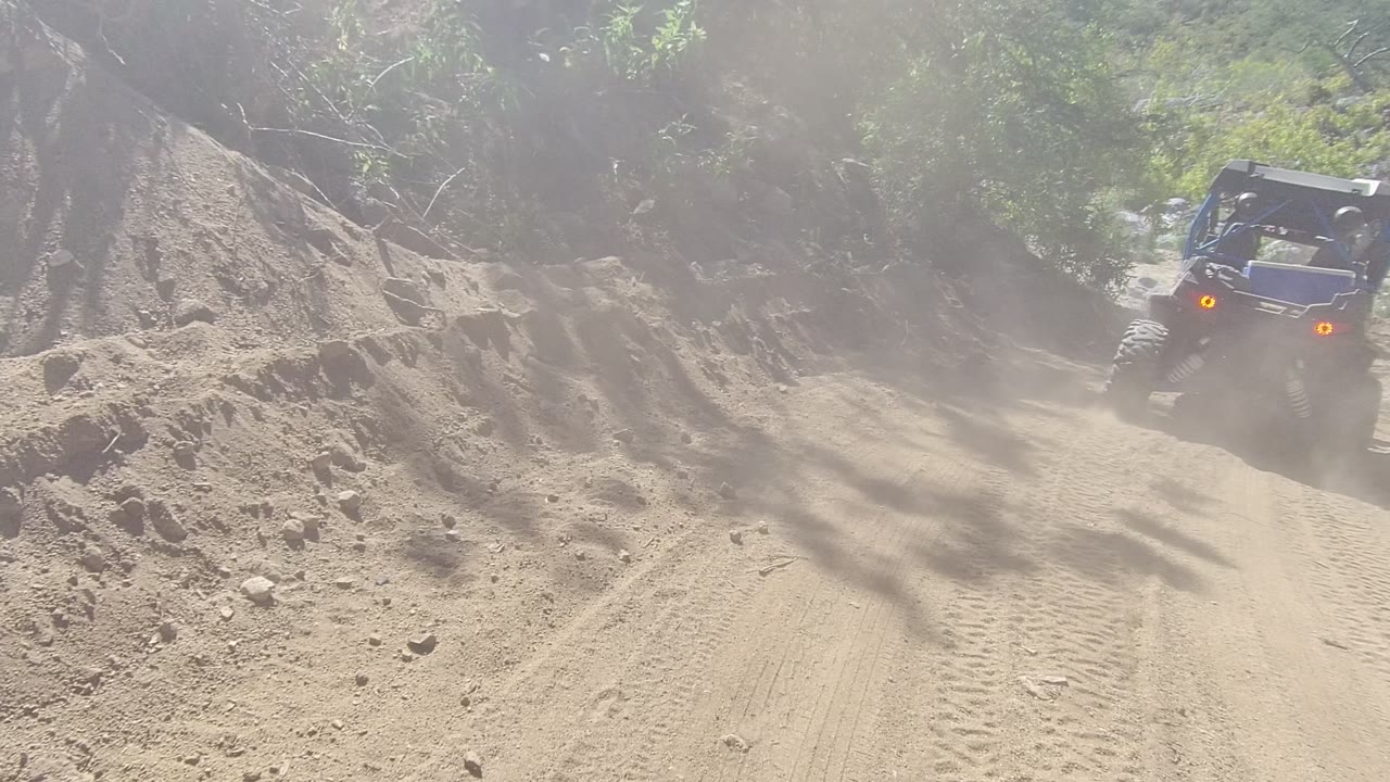 The off-road buggy reaches the right side of the frame on a dusty Los Cabos mountain trail bordered by rocks, soil, and trees, with no visible people.
