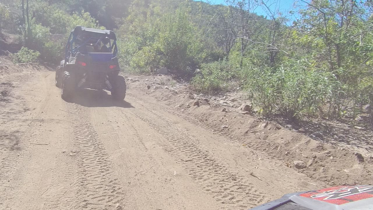 The buggy appears smaller as it travels down the Los Cabos dirt road, with a bright sky, scrubby green plants, rocky banks, and no visible people.
