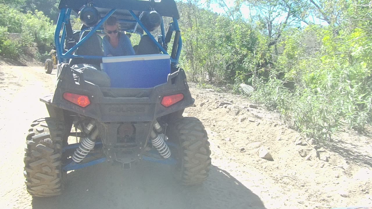 Rear view of a blue UTV close to camera on a bright dirt trail in Los Cabos, Baja California Sur, with brush and trees along the path and passengers only partly visible inside.
