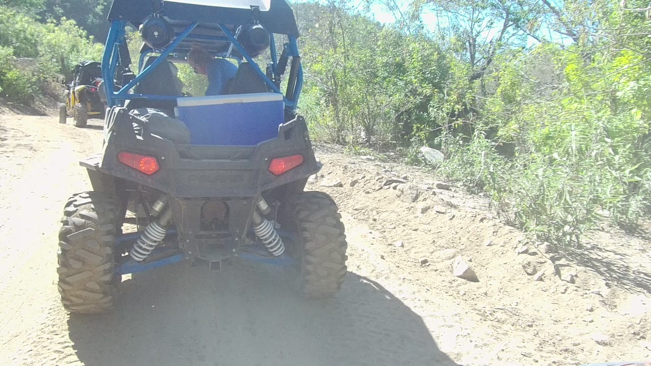 The lead blue UTV is slightly farther down the Los Cabos dirt track, framed by sunlit bushes, trees, loose rocks, and a washed-out patch of glare on the trail.