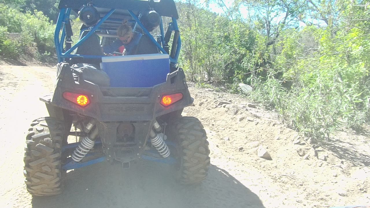 Blue off-road vehicle viewed from behind on a pale dirt trail with rocks and bushes.