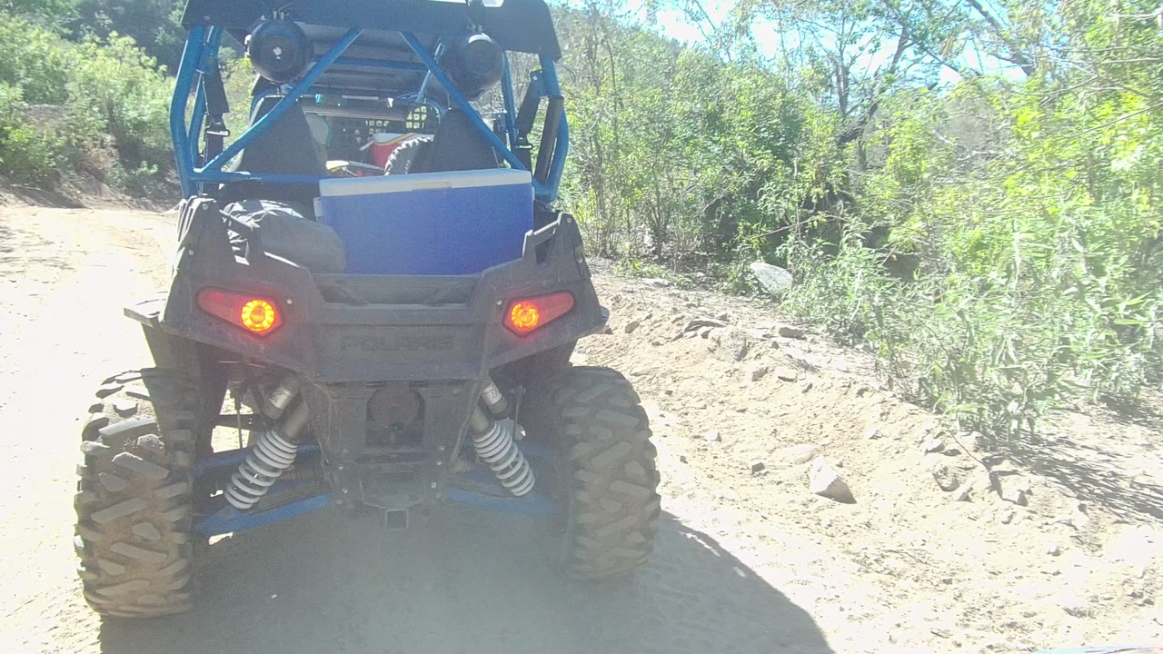 Rear view of a blue and black UTV on a dusty dirt trail in Los Cabos, with red taillights lit and bright brush along the path.
