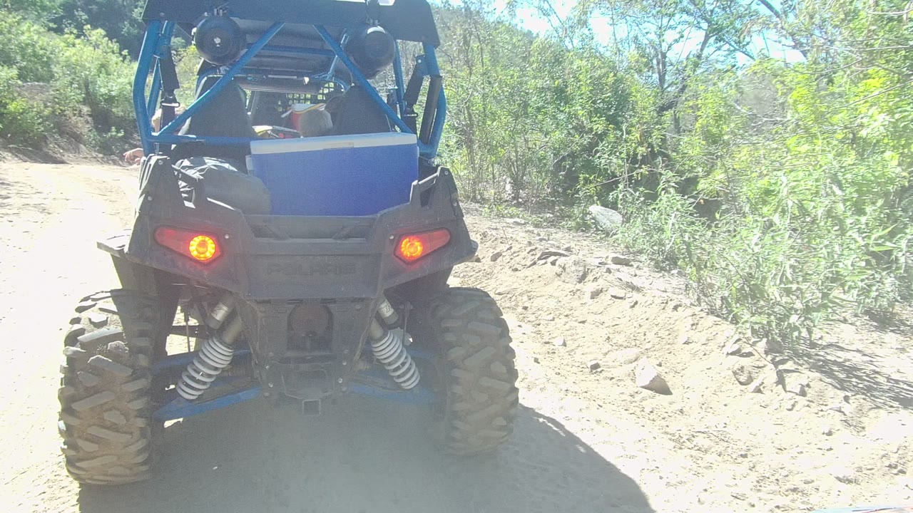 Rear view of a blue off-road vehicle stopped on a dirt trail in Los Cabos, Baja California Sur, with red taillights, rear suspension, and no visible people.